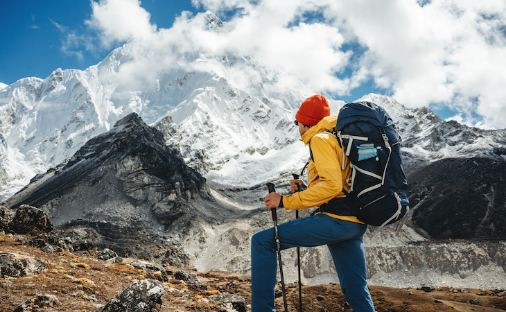 Solo tourist with travel backpack stand in front of snowy and cloudy mountains. Traveler among high altitude mountain
